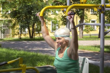 İnce, gri saçlı yaşlı bir kadın şehir parkındaki bir simülatörde bireysel fitness dersleri veriyor. Yakın plan.