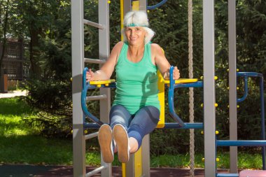 İnce, gri saçlı yaşlı bir kadın, şehir parkındaki bir simülatörde bireysel fitness derslerinde baskı yapıyor.