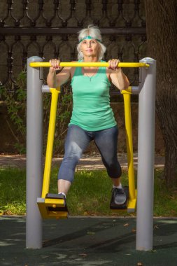 İnce, gri saçlı yaşlı bir kadın şehir parkındaki bir simülatörde bireysel fitness dersleri veriyor.