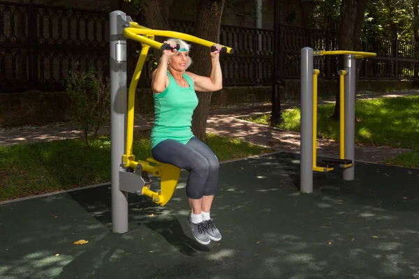 İnce, gri saçlı yaşlı bir kadın şehir parkındaki bir simülatörde bireysel fitness dersleri veriyor. Boşluğu kopyala