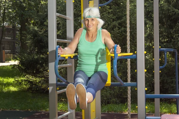 İnce, gri saçlı yaşlı bir kadın, şehir parkındaki bir simülatörde bireysel fitness derslerinde baskı yapıyor.