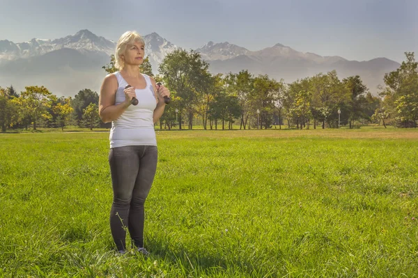 Elderly woman is engaged in fitness with a dumbbell in isolation mode outdoors in a park against a background of mountains on a sunny day