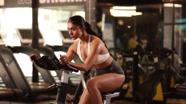 Genç bir bayan sporcu spor salonunda sabit bisiklet sürüyor, dayanıklılık ve kuvvete odaklanıyor..