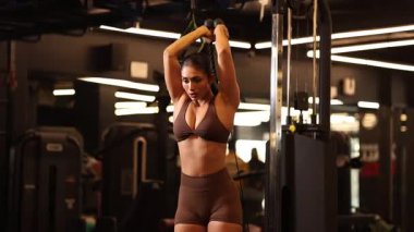 Kaslı bir kadın spor salonunda ayakta duran bir kablo triseps uzantısını güç ve hassasiyetle yapar..
