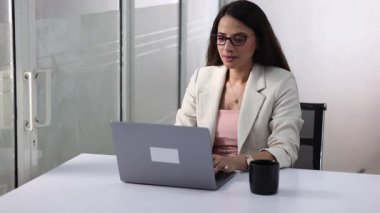 Bir kadın, parlak bir ofis alanında kahvesini yudumlarken dizüstü bilgisayarı üzerinde çalışıyor..