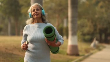 Yaşlı bir kadın, kulaklık takarak, yoga minderi taşıyarak ve dışarıda egzersiz yaparak mutlu mesut yürür..