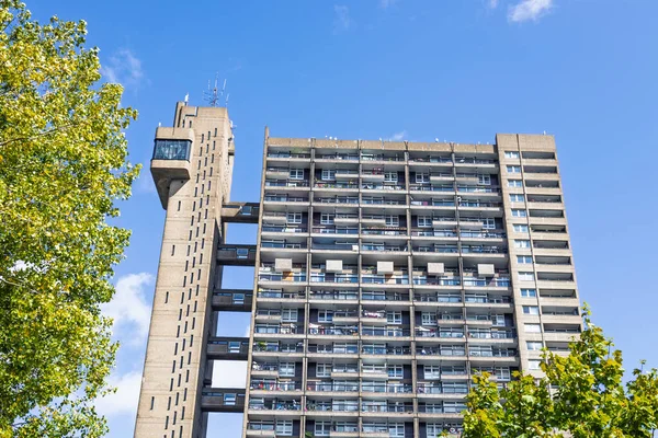 Gökdelen ve geometrik balkonları ile korunmasız beton barbar kulesi, aydınlık gün, yeşil yapraklarla çerçevelenmiş kentsel konut mimarisi.