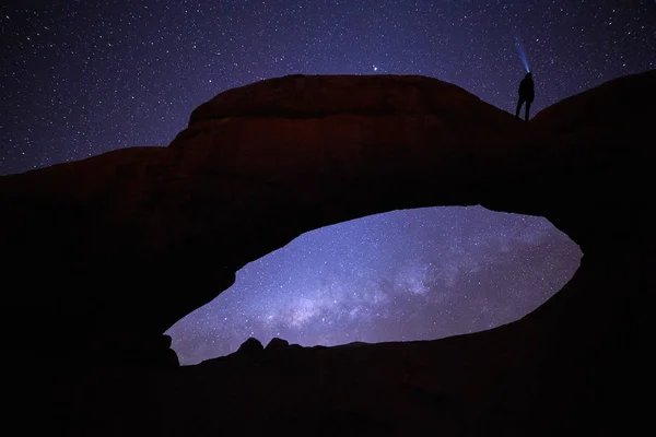 Spitzkoppe national park in Namibia. Gazing the stars on Natural Rock Arch, woman with focused beam of headlamp pointing directly to milky way. Woman silhouette under night sky. Night photography.