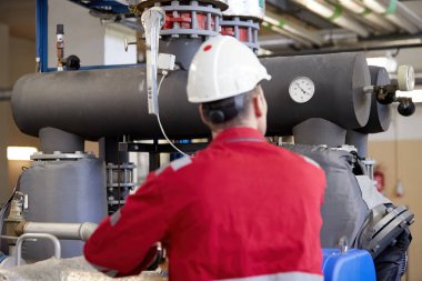 Enerji endüstrisi. Kırmızı tulum giymiş bir teknisyen ve ısıtma parametrelerini kontrol eden beyaz bir kask. Isıtma suyu dağıtım teknolojisi. Güç sektöründe hizmet çalışmaları.
