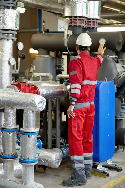 Enerji endüstrisi. Kırmızı tulum giymiş bir teknisyen ve ısıtma parametrelerini kontrol eden beyaz bir kask. Isıtma suyu dağıtım teknolojisi. Güç sektöründe hizmet çalışmaları.