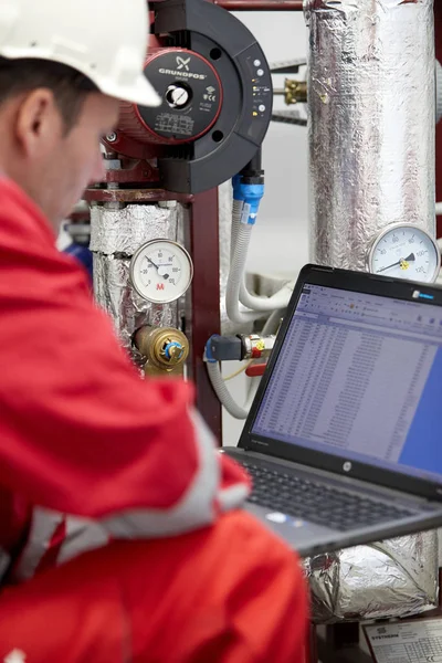 Digital control of the heating system. A technician dressed in red overalls and a white helmet check the heating parameters on laptop. Supervisory Control And Data Acquisition system in heating.