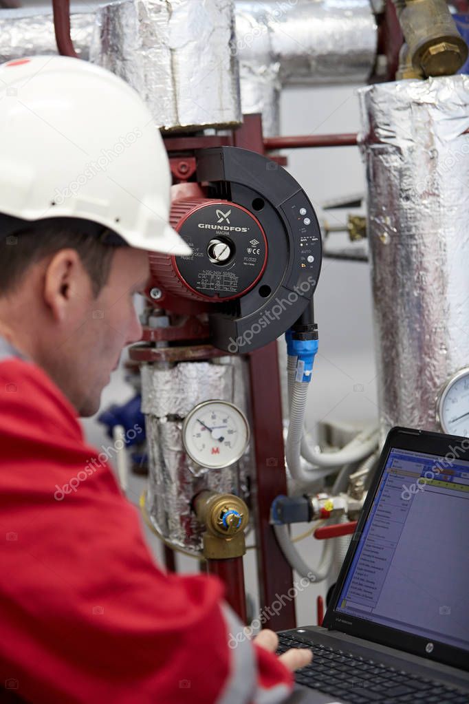 Digital control of the heating system. A technician dressed in red overalls and a white helmet check the heating parameters on laptop. Supervisory Control And Data Acquisition system in heating.