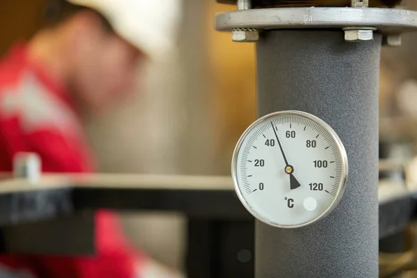 Central heating network. Thermometer on the hot water supply pipe to the heating system. Technician wearing red jacket with white helmet blurred in background. Start of the heating season.