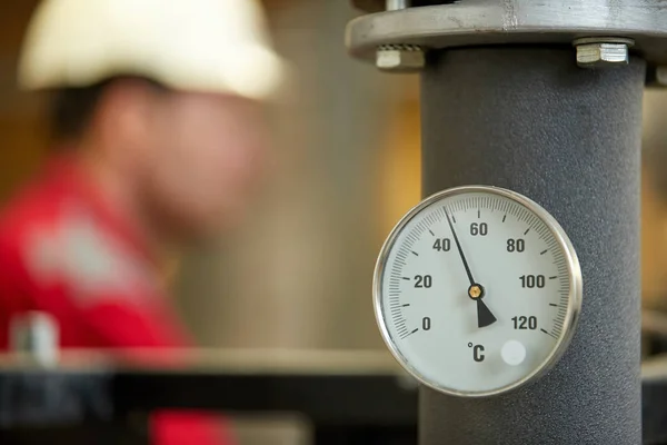 Central heating network. Thermometer on the hot water supply pipe to the heating system. Technician wearing red jacket with white helmet blurred in background. Start of the heating season.