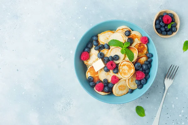 Trendy food - mini pancakes cereal. Heap of mini cereal pancakes with summer fruits in bowl on grey blue background. Copy space. Top view or flat lay.