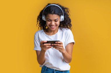 İzole edilmiş, güzel giyinmiş, güzel, harika gülüşü olan beyaz tişört, kot pantolon ve kablosuz kulaklık giyen, izin gününün tadını çıkaran akıllı telefonuyla müzik dinleyen bir kız fotoğrafı..
