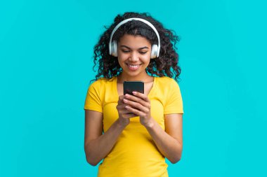 Güzel gülümseyen dak derili kız kablosuz kulaklıklarla müzik dinliyor ve akıllı telefonundaki cep telefonu uygulamasında çalma listesini tarıyor..