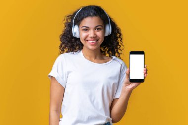 Beyaz tişörtlü, kablosuz kulaklıklı, kamerada akıllı telefonunun boş ekranını gösteren tatlı, koyu tenli bir kız. Yapılandır, metin, logo veya arayüz için boşluk kopyala.
