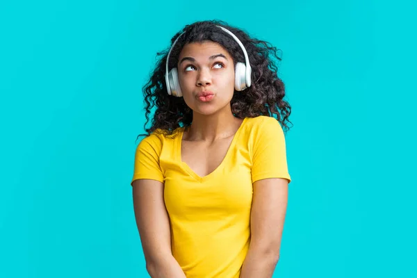 Kablosuz kulaklıklarla gülümseyen, tatlı, koyu tenli kız müzik dinliyor ve ıslık çalıyor, mavi arka planda izole edilmiş..