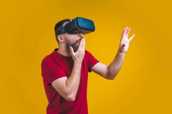 Young bearded man in basic red t-shirt and virtual reality glasses with amazed face expression touching air, isolated over bright yellow background.