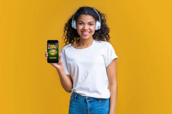 Smiling cute dark skinned girl in wireless earphones showing her phone screen to the camera with bookmaker\'s website interface. Young woman watching soccer play live broadcast online and making bets.