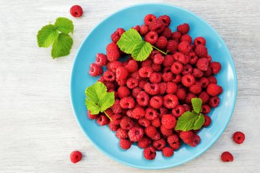 Beyaz ahşap arka planda kırmızı taze ahududu. Ahşap masa üzerinde yaprakları olan doğal organik ahududulu mavi kase; üst görünüm