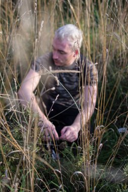 Yan görünümü alan çim bitkileri arka plan üzerinde oturan ve uzak arıyorum Genç üzgün bir çekici sakallı sarışın adam portresi.