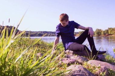 Mavi gömlek giymiş ve taş nehir arka plan üzerinde oturan ve aşağı bakıyor üzgün bir genç sıska adam.
