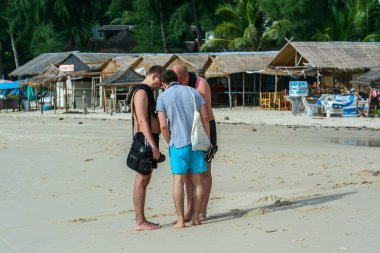 2011.05.02, Phuket, Tayland. Avrupalı turistlerin üçü sahilde durup telefon izliyor.