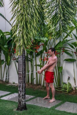 2011.05.06, Phuket, Tayland. Kırmızı yüzme şort giyen genç bir adam villanın bahçesinde bir bitkiden su hortumlama, bulanıklık etkisi.