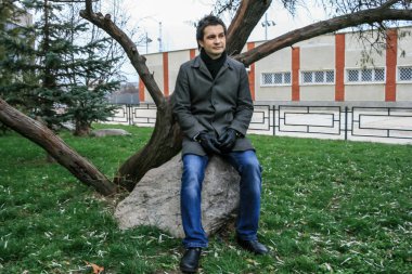 2010.11.19, Maloyaroslavets, Rusya. Parkta taş üzerinde oturan gri ceket ve kot pantolon giyen genç bir adam.