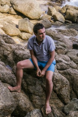 2011.05.08, Phuket, Tayland. Okyanus dalgalarının arka planında kayaların üzerinde oturan genç bir adam. Asya Turizm.