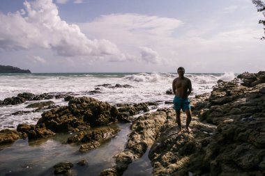 2011.05.08, Phuket, Tayland. Okyanus dalgalarının arka planında kayaların üzerinde duran genç bir adam. Asya'nın aktif turizmi.