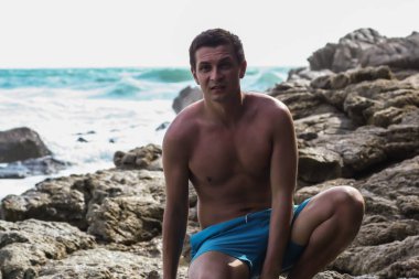 2011.05.08, Phuket, Tayland. Okyanus dalgalarının arka planında kayaların üzerinde oturan genç bir adam. Asya Turizm.