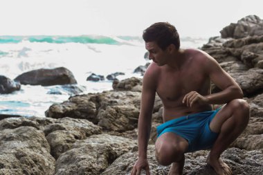 2011.05.08, Phuket, Tayland. Okyanus dalgalarının arka planında kayaların üzerinde oturan genç bir adam. Asya Turizm.