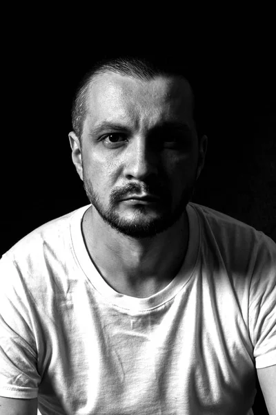 2019.05.19, Moscow, Russia. Portrait of a young serious man sitting on black background and looking to camera. Man\'s black and white portrait in the dark.