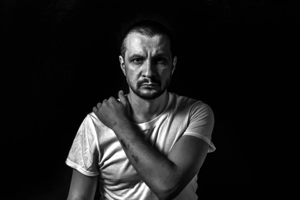 2019.05.19, Moscow, Russia. Portrait of a young serious man wearing white t-shirt sitting on black background looking to camera. Man's black and white portrait in the dark. A hand on the shoulder.