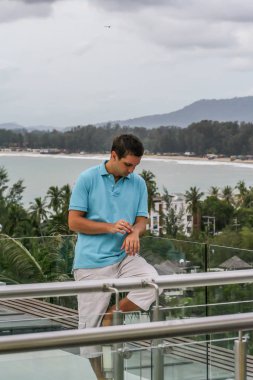 2011.05.10, Phuket, Tayland. Otelin çatısında cam çitlerin yanında duran genç bir adam. Phuket plajının arka planında kameraya bakan bir adam.