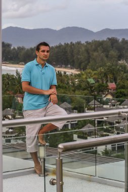 2011.05.10, Phuket, Tayland. Otelin çatısında cam çitlerin yanında duran genç bir adam. Phuket plajının arka planında kameraya bakan bir adam.