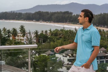 2011.05.10, Phuket, Tayland. Otelin çatısında cam çitlerin yanında duran genç bir adam. Phuket plajının arka planında kameraya bakan bir adam.