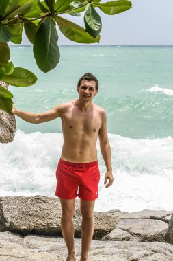 2011.05.10, Phuket, Tayland. Kırmızı yüzme şortu giyen genç bir adam kayaların üzerinde durup okyanusa bakıyor. Yaz sahilde istirahat. 