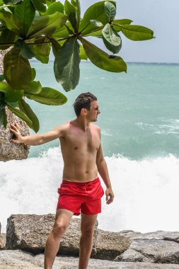 2011.05.10, Phuket, Tayland. Kırmızı yüzme şortu giyen genç bir adam kayaların üzerinde durup okyanusa bakıyor. Yaz sahilde istirahat. 
