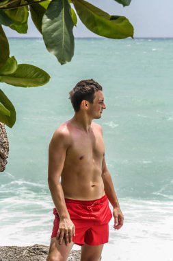 2011.05.10, Phuket, Tayland. Kırmızı yüzme şortu giyen genç bir adam kayaların üzerinde durup okyanusa bakıyor. Yaz sahilde istirahat. 