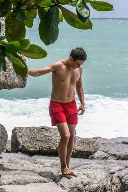 2011.05.10, Phuket, Tayland. Kırmızı yüzme şortu giyen genç bir adam spor ayakkabılarını çıkarıyor. Kayalıklarda bir adam..