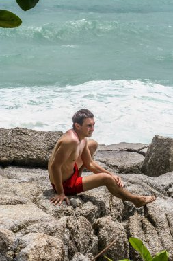 2011.05.10, Phuket, Tayland. Kırmızı yüzme şortu giyen genç bir adam kayaların üzerinde oturmuş okyanusa bakıyor. Yaz sahilde istirahat. 