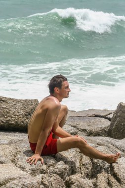 2011.05.10, Phuket, Tayland. Kırmızı yüzme şortu giyen genç bir adam kayaların üzerinde oturmuş okyanusa bakıyor. Yaz sahilde istirahat. 