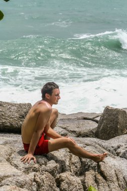 2011.05.10, Phuket, Tayland. Kırmızı yüzme şortu giyen genç bir adam kayaların üzerinde oturmuş okyanusa bakıyor. Yaz sahilde istirahat. 