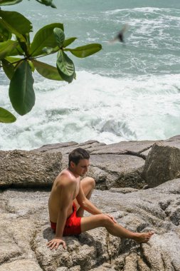 2011.05.10, Phuket, Tayland. Kırmızı yüzme şortu giyen genç bir adam kayaların üzerinde oturmuş okyanusa bakıyor. Yaz sahilde istirahat. 