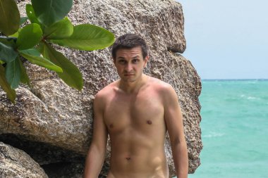 2011.05.10, Phuket, Tayland. Okyanus kıyısındakayanın arka planında duran üstsüz genç bir adam..