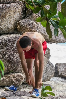 2011.05.10, Phuket, Tayland. Kırmızı yüzme şortu giyen genç bir adam spor ayakkabılarını çıkarıyor. Kayalıklarda bir adam..